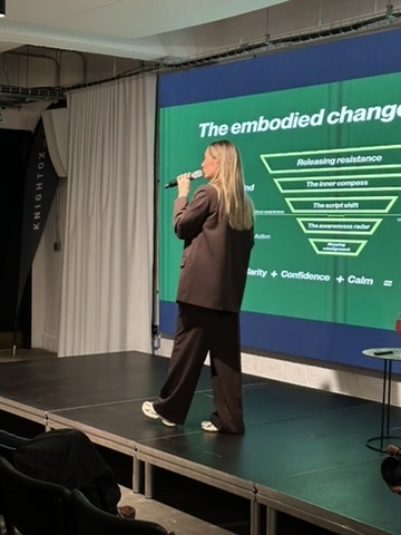 Lucy presenting on stage with a microphone, a slide on the screen behind her